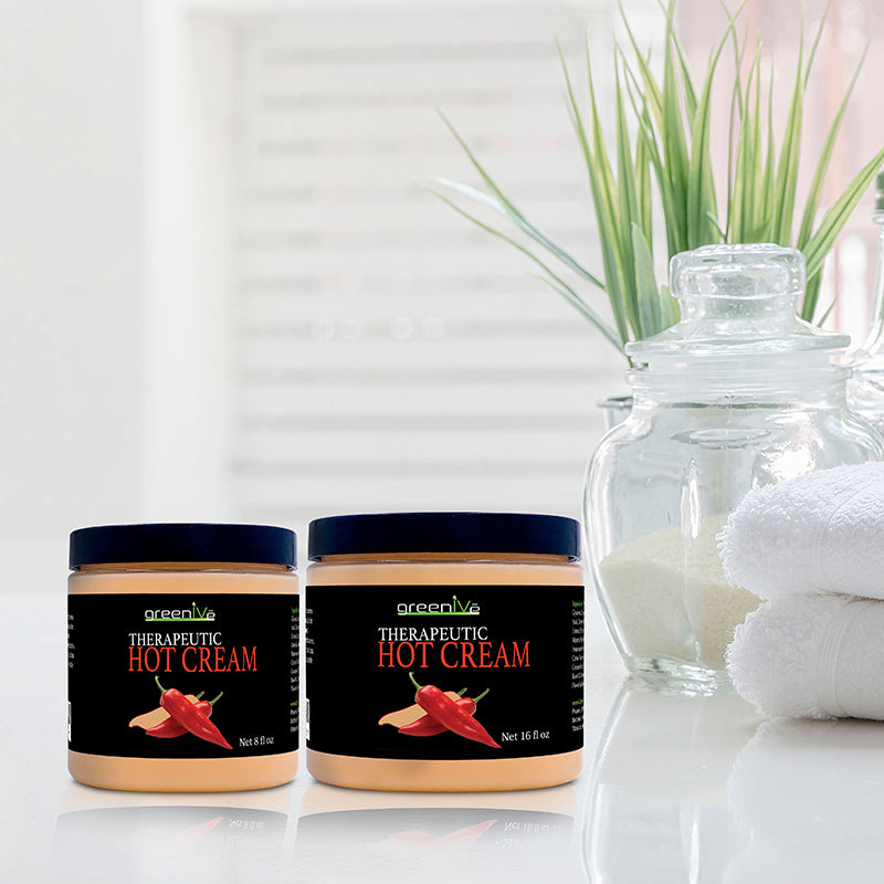 Two jars of Therapeutic Hot Cream on a reflective surface with a blurred plant and towels in the background.