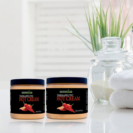 Two jars of Therapeutic Hot Cream on a reflective surface with a blurred plant and towels in the background.
